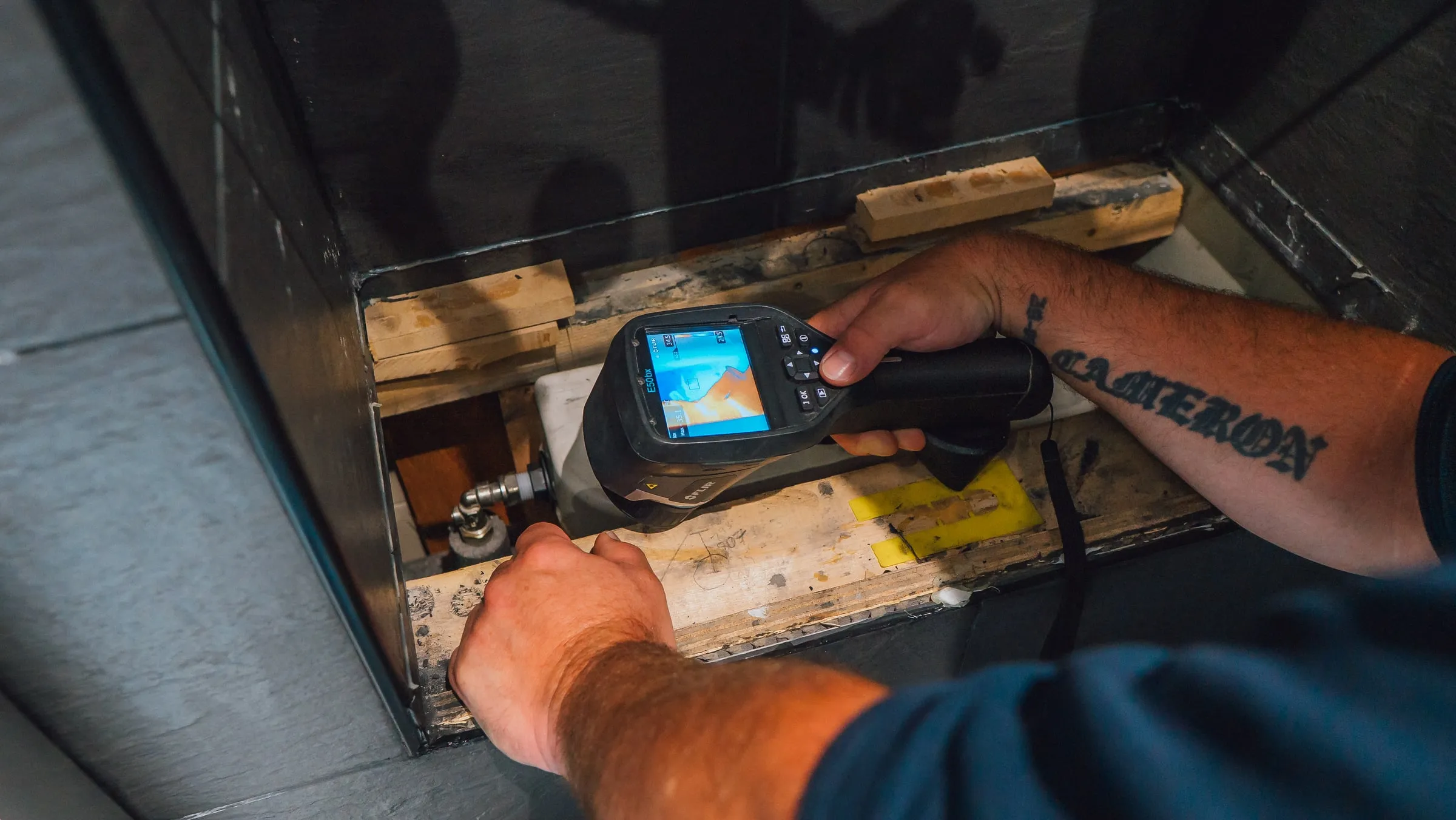Engineer holding a thermal camera into an opened bathroom cabinet panel beside a pipe valve, the screen showing the heat pattern