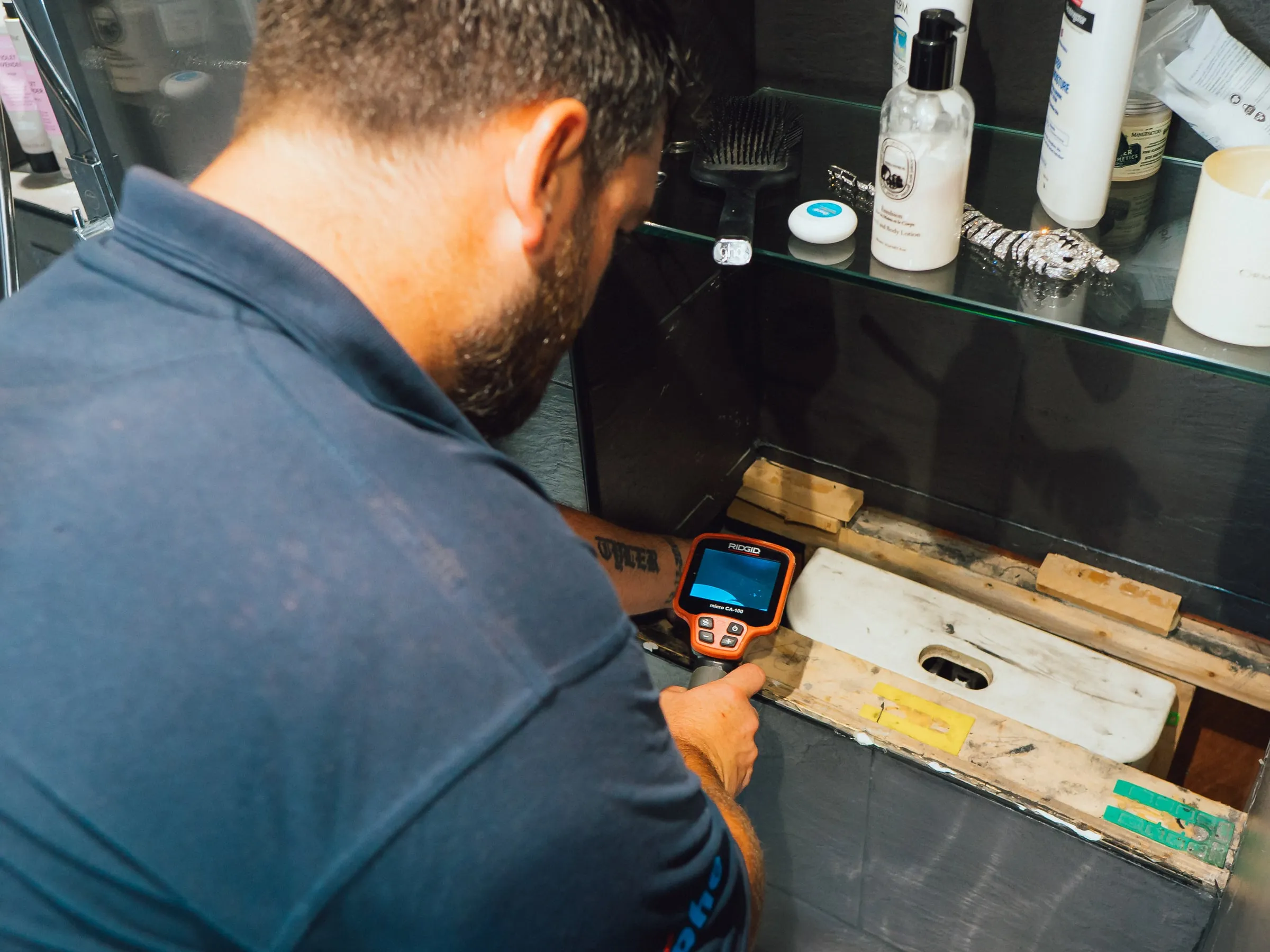 Engineer checking the screen of a thermal camera held to the opened base of a bathroom cabinet, toiletries on the glass shelf above