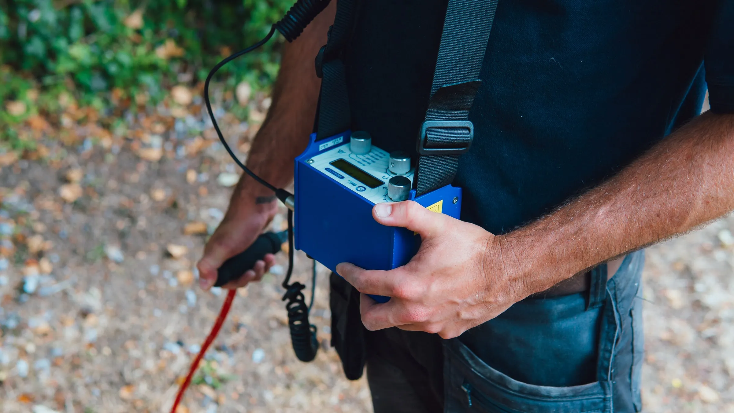Engineer holding a blue leak detection unit on a shoulder strap, its sensor cable in his other hand