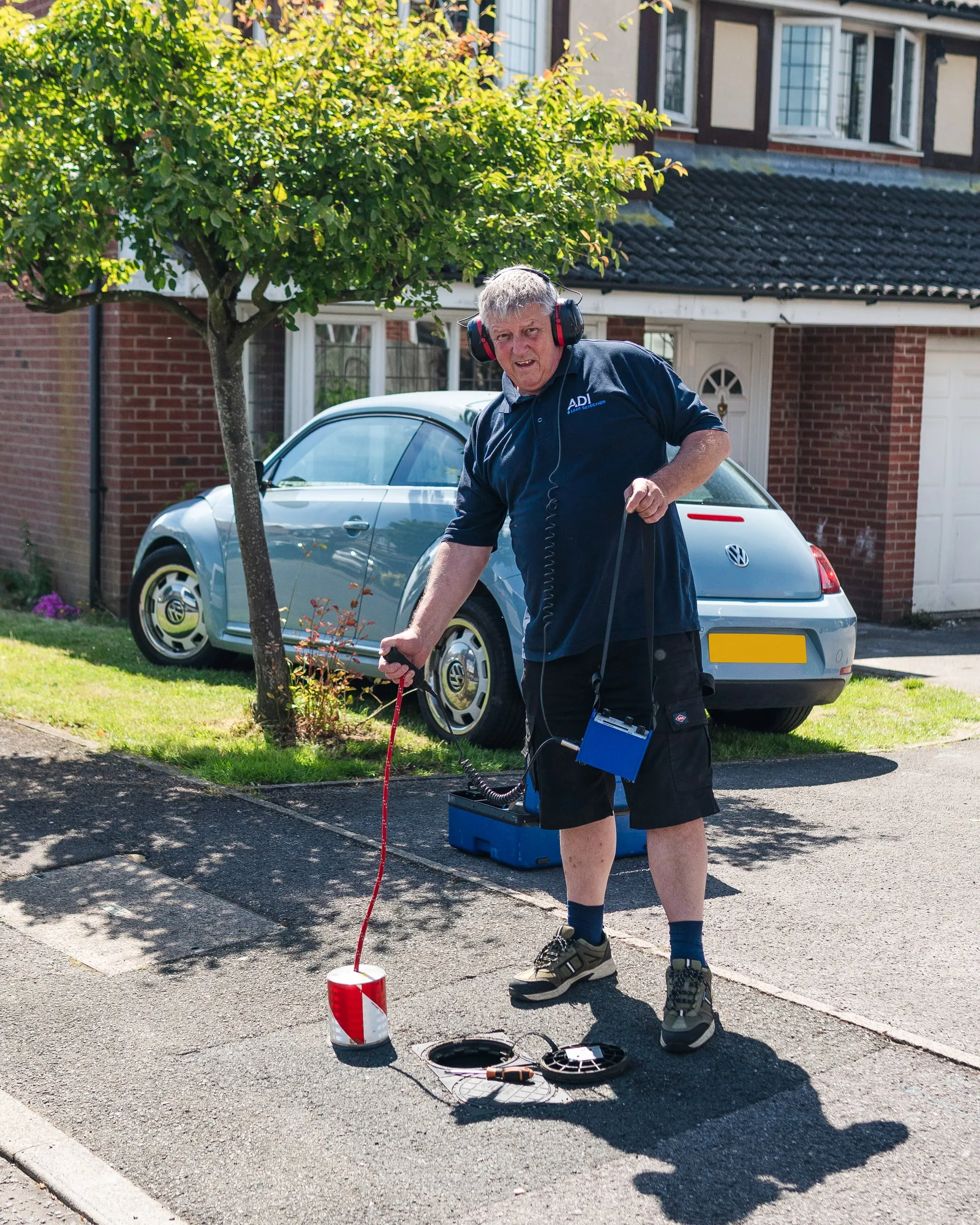 Engineer wearing ear defenders listening with a ground microphone on a residential driveway, beside an open stopcock chamber
