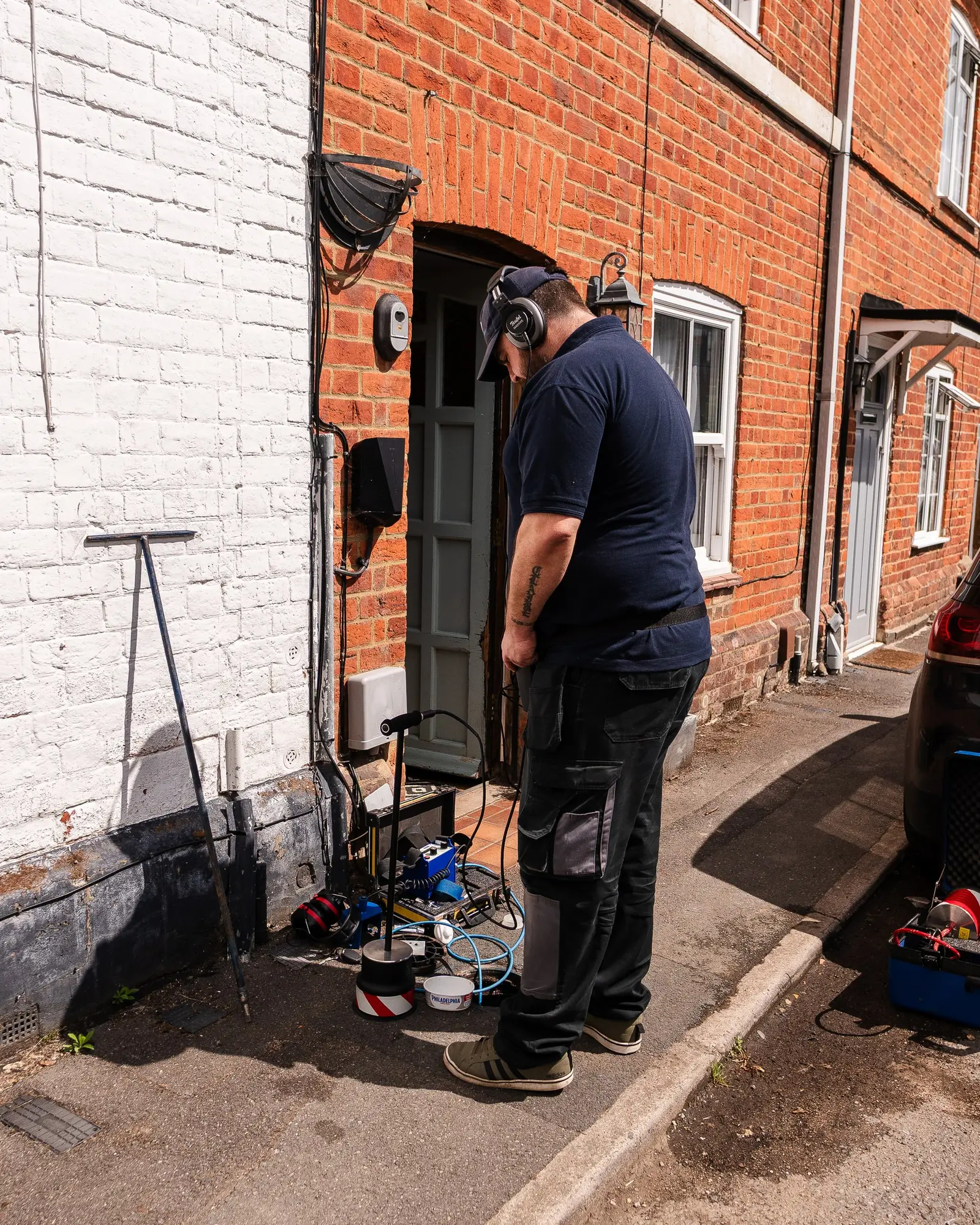 ADI engineer with ear defenders at the open front door of a red-brick terraced house, leak detection equipment on the pavement beside him