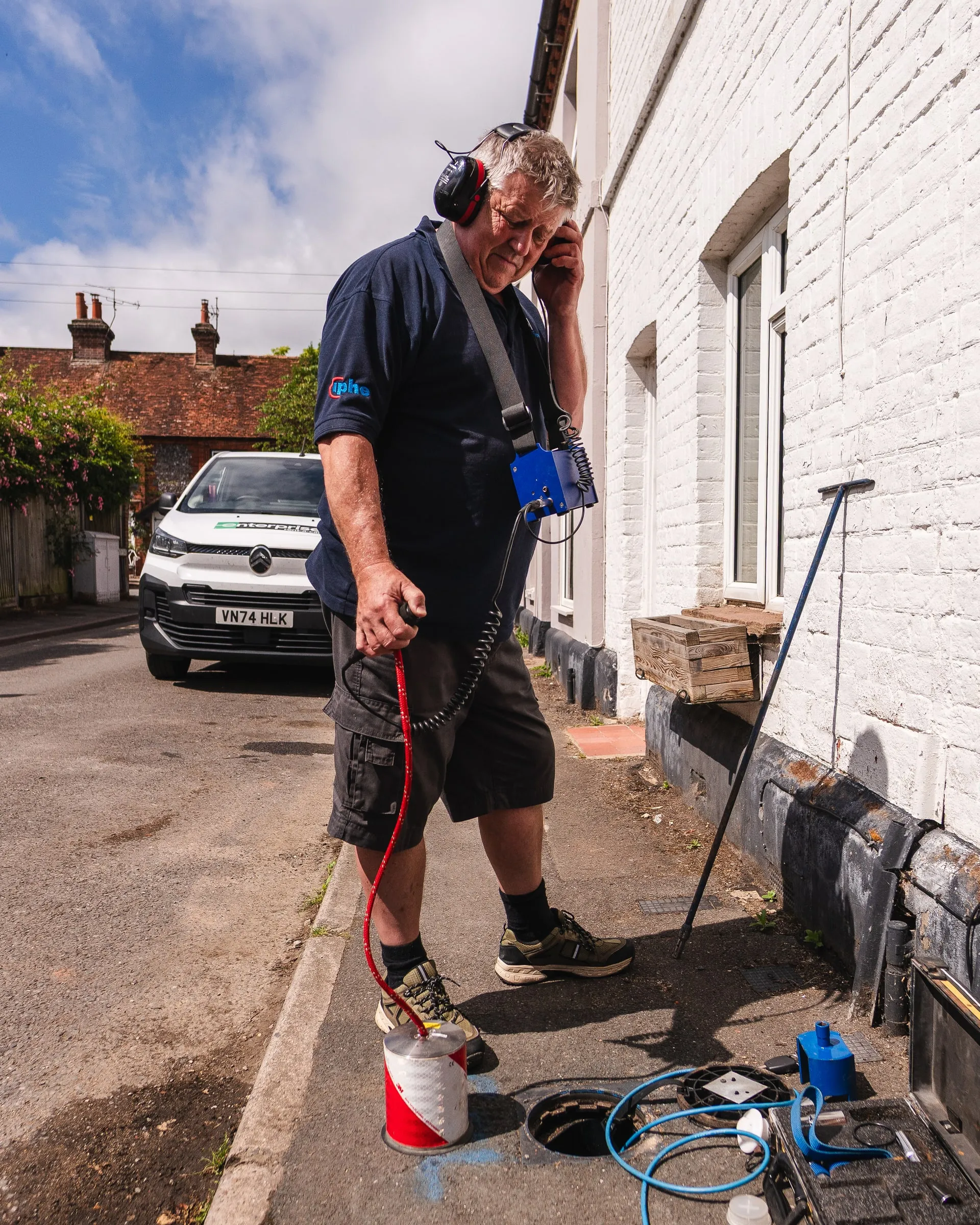 Engineer wearing ear defenders listening with a ground microphone along the pavement outside a white-painted terraced house